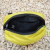Top-down view of an open yellow-green zippered pouch with black interior lining, an internal zip pocket and key clip, resting on pale gravel.