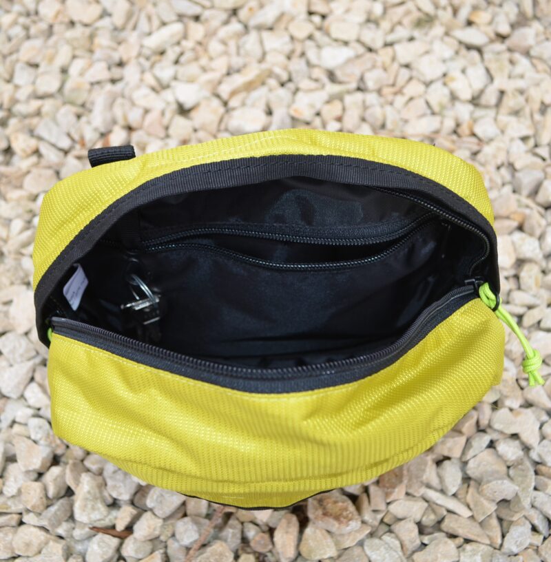 Top-down view of an open yellow-green zippered pouch with black interior lining, an internal zip pocket and key clip, resting on pale gravel.