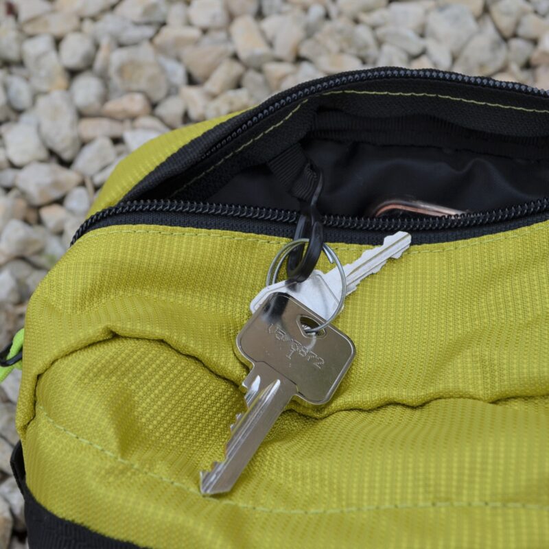 Silver house key on a small keyring lies on a partially open lime-green zippered pouch, with a smaller key and black zipper visible against pale gravel.