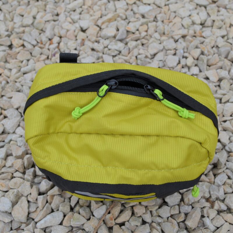 Top-down view of a small lime-green ripstop zippered pouch with black trim and neon-green cord zipper pulls resting on pale gravel stones.