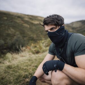 A man in a short-sleeved green top, black neck gaiter and gloves crouches on a grassy hillside, gazing to the side with rolling hills and a cloudy sky behind him.