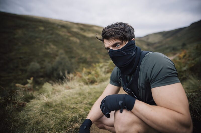 A man in a short-sleeved green top, black neck gaiter and gloves crouches on a grassy hillside, gazing to the side with rolling hills and a cloudy sky behind him.