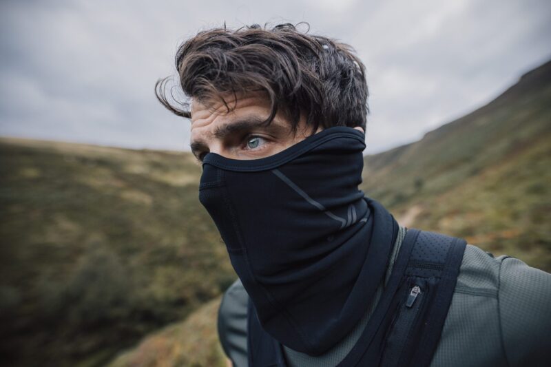 Close-up portrait of a man with tousled brown hair and blue eyes wearing a black neck gaiter and outdoor gear on a windswept grassy hillside under a cloudy sky
