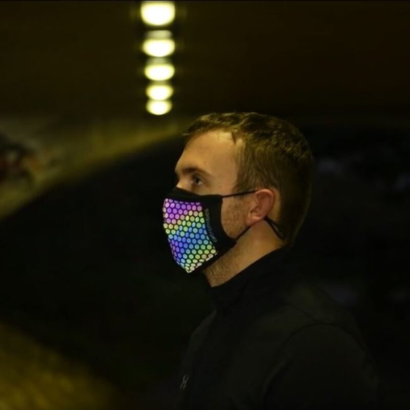 Profile of a man standing in a dim tunnel wearing a black face mask covered in a multicoloured hexagon LED pattern, with a row of glowing overhead lights.