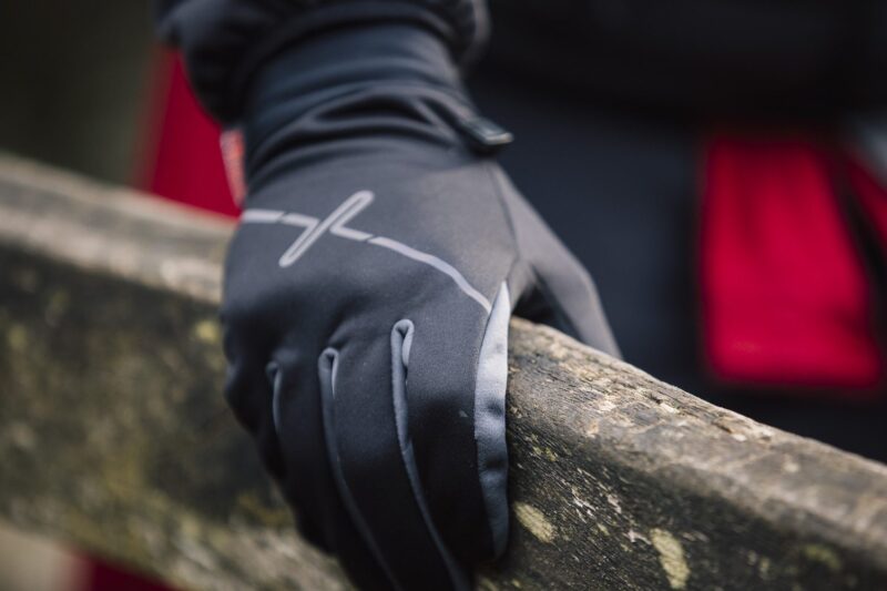 Close-up of a black insulated glove with a reflective stripe gripping a weathered wooden railing, with a blurred red coat in the background.