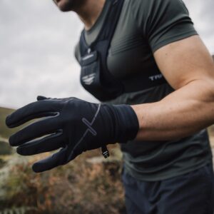 Runner on a hillside trail wearing black performance gloves and a grey-green short-sleeve top, leaning forward with a gloved hand extended toward the camera against moorland and a cloudy sky.