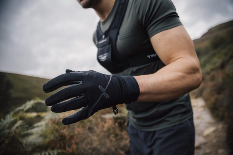 Runner on a hillside trail wearing black performance gloves and a grey-green short-sleeve top, leaning forward with a gloved hand extended toward the camera against moorland and a cloudy sky.