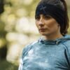 Woman with brown hair in a ponytail wearing a black headband and blue-grey athletic hoodie, standing outdoors with a neutral expression against a soft, sunlit bokeh background.