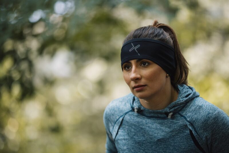 A woman with her hair in a ponytail wearing a black headband with a grey logo and a blue heathered hoodie leans forward with a focused expression against a softly blurred leafy outdoor background.