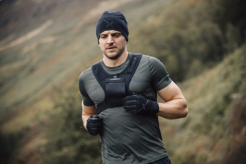 A man with a short beard runs through a misty, grassy hillside wearing a black beanie, fitted grey-green training top, a front chest vest and black gloves.