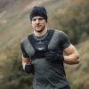 Man wearing a black beanie, fitted green training shirt, chest-weighted vest labelled 'FREE TRAIN' and black gloves running outdoors on a blurred hillside landscape.