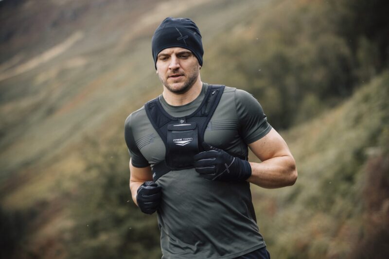 Man wearing a black beanie, fitted green training shirt, chest-weighted vest labelled 'FREE TRAIN' and black gloves running outdoors on a blurred hillside landscape.