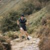 Man in a beanie, gloves and a weighted vest running uphill mid-stride on a rocky country trail through ferns and rolling grassy hills.
