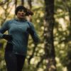 Woman jogging through a leafy forest wearing a teal long-sleeve top, black headband and gloves, her hair swept back with trees softly blurred behind her