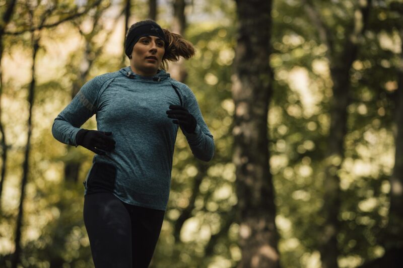 Woman jogging through a leafy forest wearing a teal long-sleeve top, black headband and gloves, her hair swept back with trees softly blurred behind her