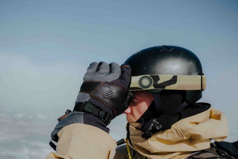 Close-up of a person in a beige winter jacket wearing a black helmet with a camouflage goggle strap, lifting a grey-and-black glove to adjust goggles against a pale blue sky and snowy background.