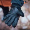 Close-up of an outstretched black leather winter glove with orange stitching and an insulated cuff with a drawcord, held against a blurred rocky outdoor background.