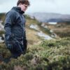 A person wearing a dark waterproof jacket, gloves and glasses walks through low green heath and rocky outcrops with a distant loch under an overcast sky.