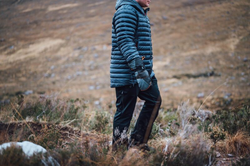 Person wearing a blue puffer jacket, mittens and dark trousers walking across windswept moorland with brown grasses and low shrubs.