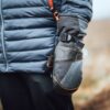 Close-up of a person wearing a blue‑grey puffer jacket and grey insulated mittens with black leather patches, orange stitching and a buckle strap, standing outdoors against a blurred brown landscape.