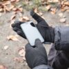 Hands in black touchscreen gloves holding a smartphone with a blank screen above leaf-strewn ground, a dark winter jacket sleeve visible at the wrist.
