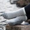 Feet in thick light-grey ribbed socks with dark-grey reinforced toes and heels, legs in black leggings, resting on a rock beside a blurred flowing stream.