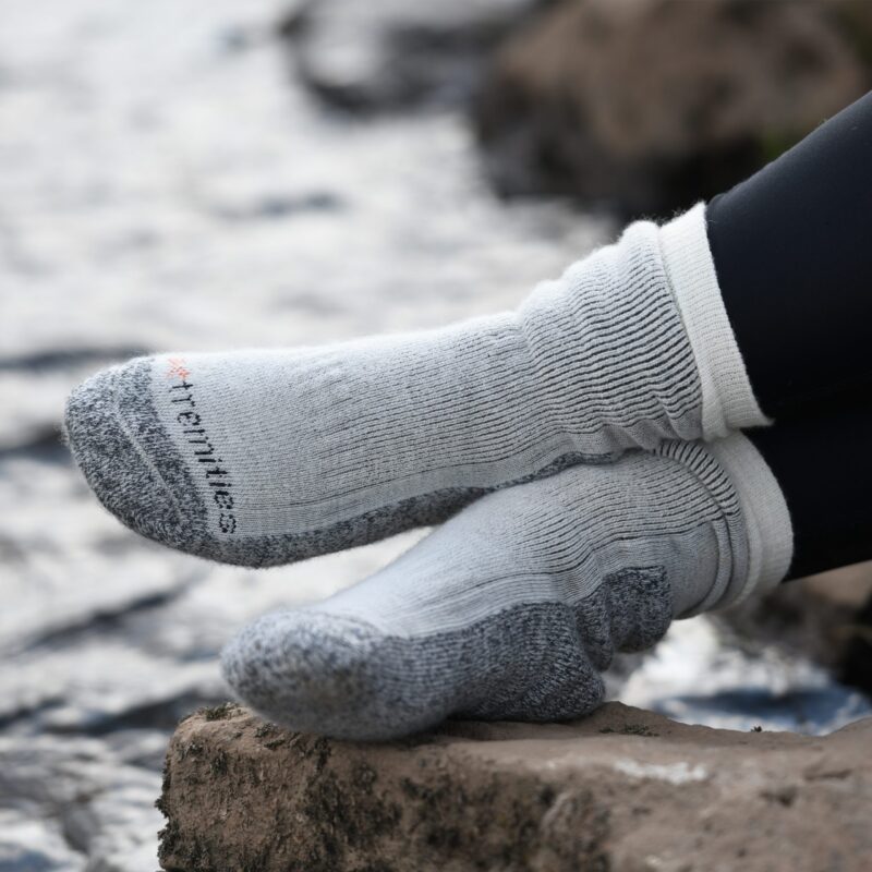 Feet in thick light-grey ribbed socks with dark-grey reinforced toes and heels, legs in black leggings, resting on a rock beside a blurred flowing stream.