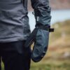 Close-up of a person's side wearing a dark waterproof jacket and a black Gore-Tex mitten with an adjustable wrist strap, set against a blurred outdoor landscape.