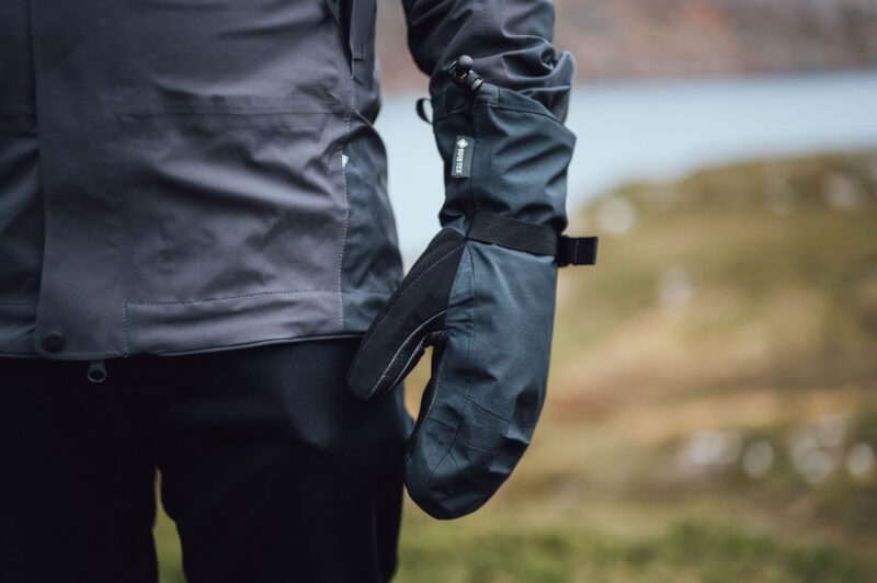 Close-up of a person's side wearing a dark waterproof jacket and a black Gore-Tex mitten with an adjustable wrist strap, set against a blurred outdoor landscape.