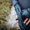 Close-up of a person wearing dark puffy mittens adjusting a strap on a black hand cover while holding hiking poles with cork handles against a blurred green ground background.