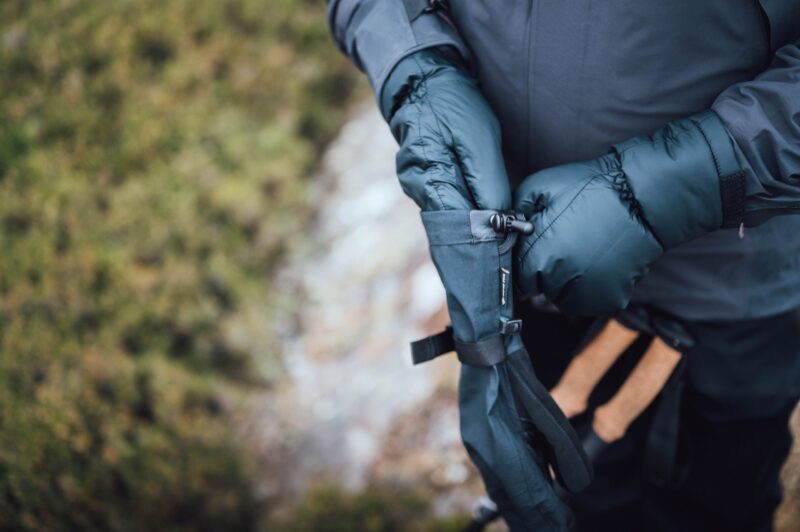 Close-up of a person wearing dark puffy mittens adjusting a strap on a black hand cover while holding hiking poles with cork handles against a blurred green ground background.