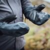 Close-up of a person wearing black heavy-duty mitten-style waterproof gloves with leather palms and adjustable wrist straps, held out against a blurred outdoor landscape