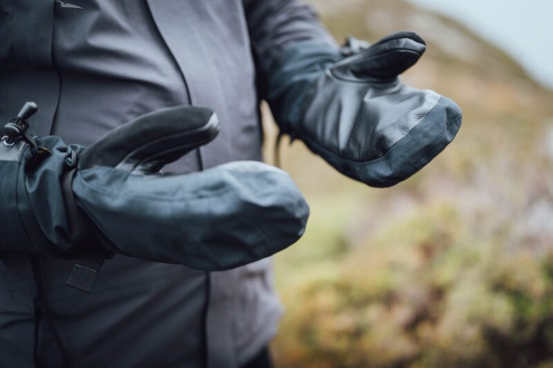 Close-up of a person wearing black heavy-duty mitten-style waterproof gloves with leather palms and adjustable wrist straps, held out against a blurred outdoor landscape