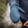 Close-up of an outstretched padded teal mitten showing visible stitching, worn with a dark jacket against a blurred outdoor background.