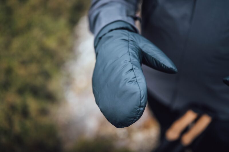 Close-up of an outstretched padded teal mitten showing visible stitching, worn with a dark jacket against a blurred outdoor background.