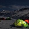 Cluster of glowing green and red tents pitched on a snow-covered mountainside at night beneath a clear, starry sky and distant snow-capped peaks