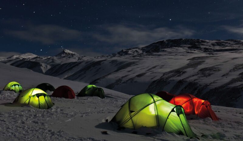 Cluster of glowing green and red tents pitched on a snow-covered mountainside at night beneath a clear, starry sky and distant snow-capped peaks