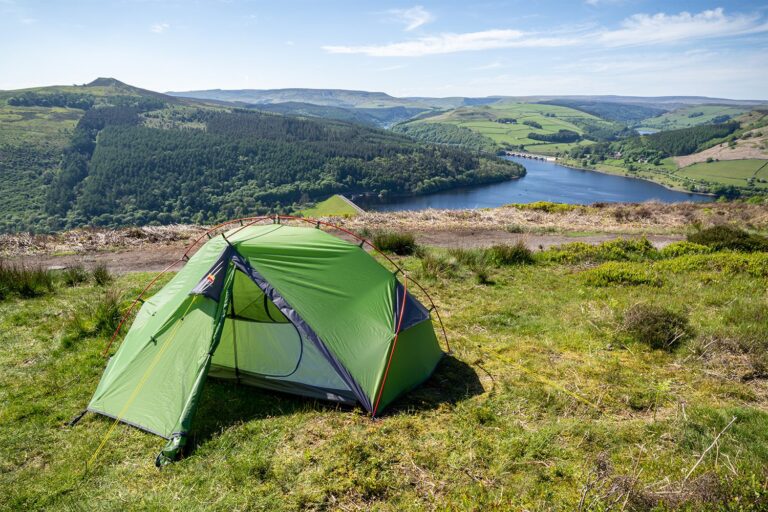 Bright green dome tent pitched on a grassy hillside overlooking a tree-covered valley and a blue reservoir with a stone viaduct and rolling hills under a clear sky