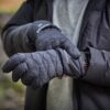 Close-up of a person outdoors wearing quilted black 'extremities' insulated gloves and a dark puffer jacket, one glove adjusting a smartwatch on the other wrist against a blurred stone-wall background.