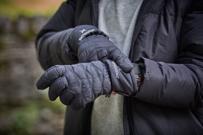 Close-up of a person outdoors wearing quilted black 'extremities' insulated gloves and a dark puffer jacket, one glove adjusting a smartwatch on the other wrist against a blurred stone-wall background.