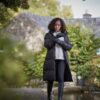 Woman in a long black puffer coat, grey sweater and gloves adjusts her sleeve while walking along a path by a stream, with a stone cottage and autumnal greenery in the background.