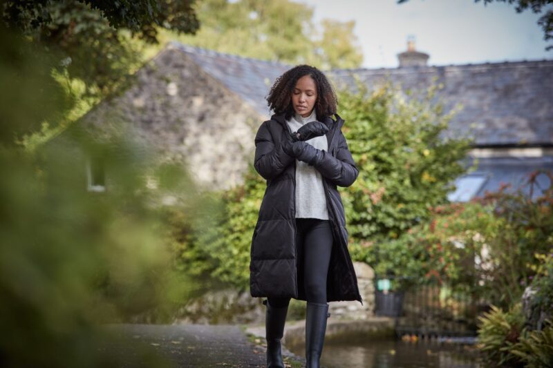 Woman in a long black puffer coat, grey sweater and gloves adjusts her sleeve while walking along a path by a stream, with a stone cottage and autumnal greenery in the background.