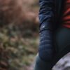 Close-up of a person outdoors wearing navy quilted mittens with an "extremities" logo, a red zip-up jacket and dark leggings with one knee bent against a blurred woodland background.