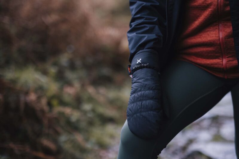 Close-up of a person outdoors wearing navy quilted mittens with an "extremities" logo, a red zip-up jacket and dark leggings with one knee bent against a blurred woodland background.
