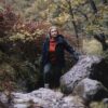Woman in a dark waterproof jacket and orange fleece climbing over mossy rocks in an autumnal woodland, steadying herself with a gloved hand amid ferns and trees.