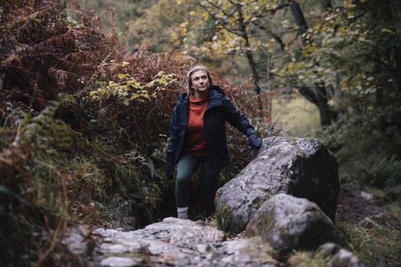 Woman in a dark waterproof jacket and orange fleece climbing over mossy rocks in an autumnal woodland, steadying herself with a gloved hand amid ferns and trees.