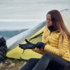 A woman in a yellow puffer jacket and black leggings sits by a small yellow-and-silver tent on a grassy hillside, putting on black gloves with a black backpack beside her and cloudy hills in the background.