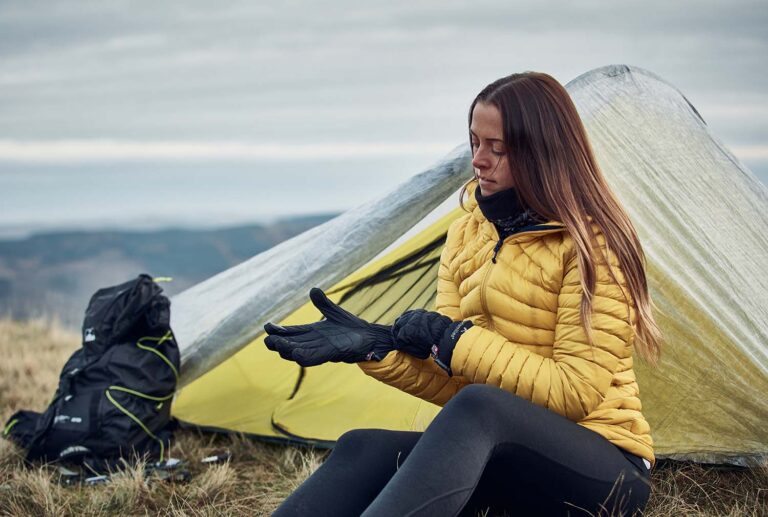 A woman in a yellow puffer jacket and black leggings sits by a small yellow-and-silver tent on a grassy hillside, putting on black gloves with a black backpack beside her and cloudy hills in the background.