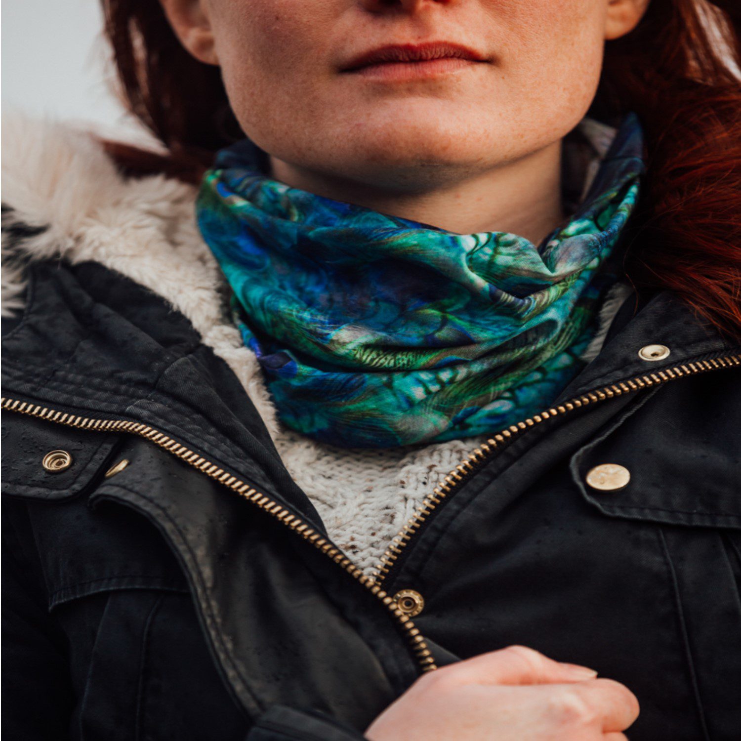 Peacock-neck-tube-lifestyle-1-1.jpg Close-up of a woman's lower face and neck wearing a blue-green patterned neck gaiter over a cream knitted jumper, a dark fur-trimmed jacket with a brass zipper and a hand gripping the jacket.
