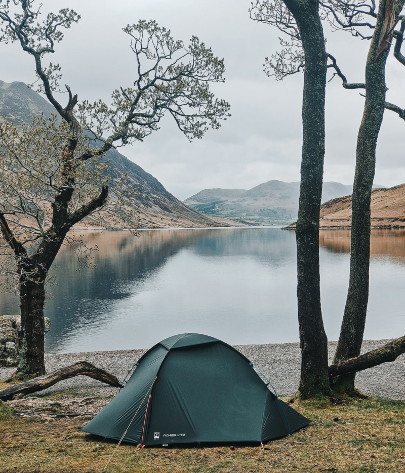 Dark green dome tent pitched on a grassy lakeshore beneath bare, twisting trees, overlooking a calm lake that mirrors surrounding hills and a cloudy sky.